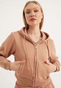 Felpa zip con cappuccio di un delicato rosa, realizzata in un tessuto morbido, con tasca frontale, cappuccio con coulisse e polsini a costine. Il modello posa con le mani nelle tasche.