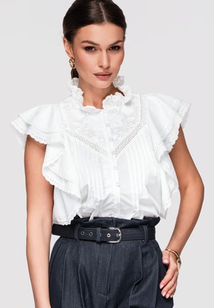 Kobieta ubrana w białą bluzkę z falbankami i kwiatowym haftem oraz ciemne spodnie z wysokim stanem, czarnym paskiem i złotą bransoletką.