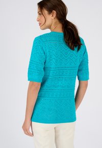 Suéter de punto en tono azul verdoso con mangas cortas, que presenta un patrón en zigzag y una textura de tejido abierto. Los puños y el dobladillo acanalados realzan el diseño.