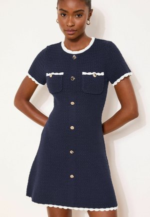 Frau trägt ein marineblaues, strukturiertes Kleid mit kurzen Ärmeln, weißer Paspelierung, vorderen Knöpfen und zwei Brusttaschen mit Knöpfen.