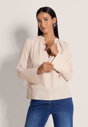 Hellbeige Bluse mit vertikalen Streifen, V-Ausschnitt, Glockenärmeln und Bändern an den Manschetten. Kombiniert mit blauen Jeans.
