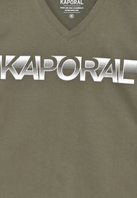 Camiseta verde oliva con escote en forma de V, presenta letras en negrita blancas de "KAPORAL" en un patrón de puntos, textura de tela suave.