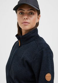 Marineblå fleecjakke med glidelås og høy krage, med en beige lærlogo på venstre arm og strukturert stoff.