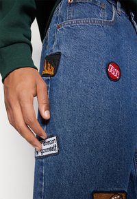 Jeans i denim med olika broderade lappar. Nyckelfärger inkluderar blå denim, svart, röd och vit. Lapparna visar text och lågor.