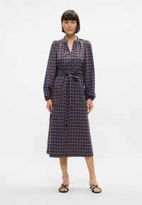 Rutet kjole i mørke nyanser med blå linjer, lange ermer, v-hals og belte i midjen. Materialet ser mykt og lett ut.