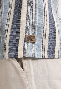 Gestreepte geweven stof in blauw, wit en beige met een gestructureerde oppervlak. Bevat een bruine label met "Petrol Ind" erop gestikt.