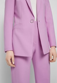 Femme portant un blazer lilas sur mesure avec un seul fermoir rond en argent et un pantalon assorti, debout devant un fond uni.