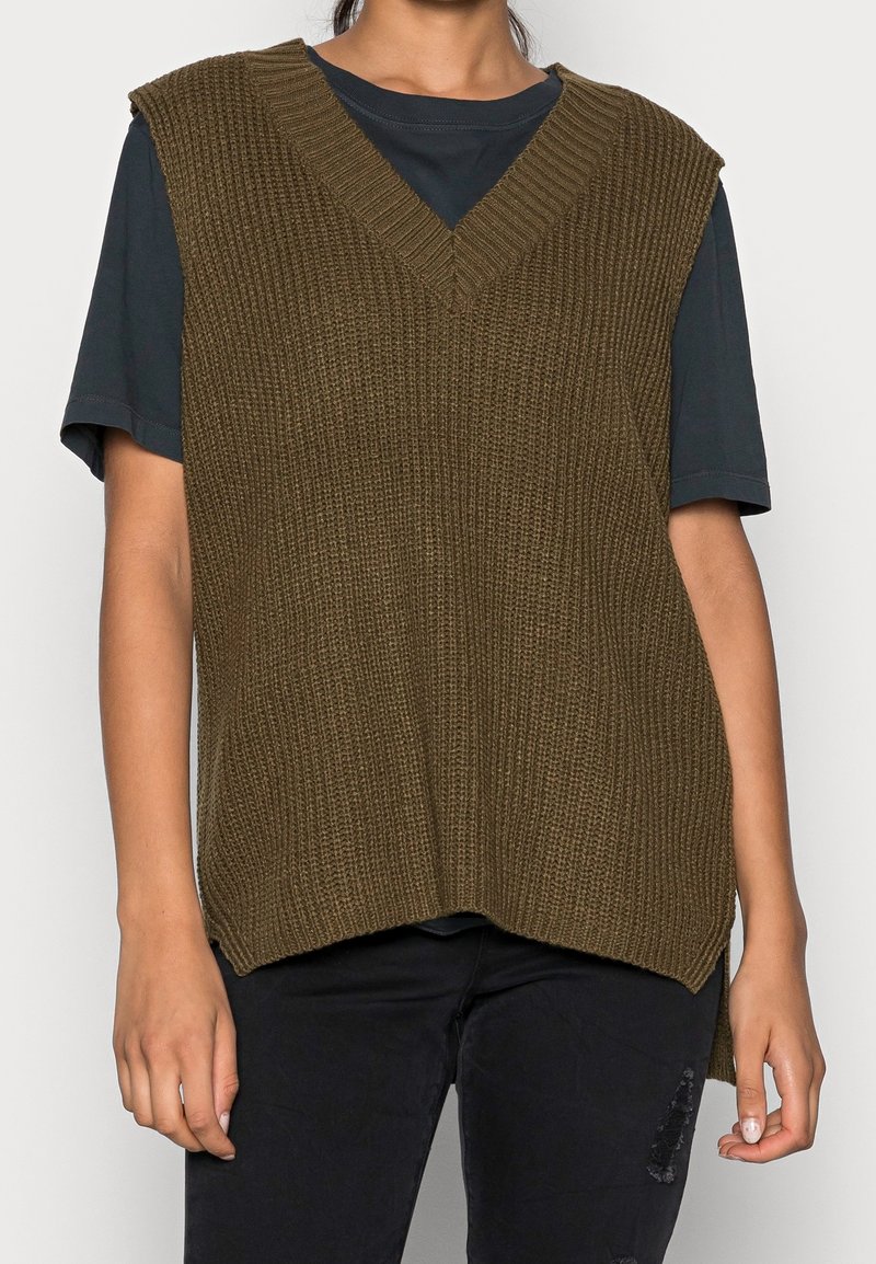 Personne portant un gilet sans manches tricoté vert olive foncé sur une chemise noire à manches courtes et un pantalon noir avec un détail déchiré.