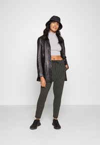 Casaco longo de couro preto, top cropped cinza claro, calças verdes listradas, chapéu tipo balde preto e ténis pretos. O conjunto combina elementos casuais e ajustados.