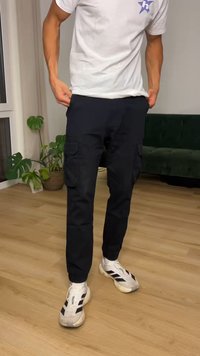 Személy fekete cargo nadrágot igazít egy fahatású padlójú szobában, fehér pólóban, fehér sportcipőben, közelében zöld kanapé és cserepes növények.