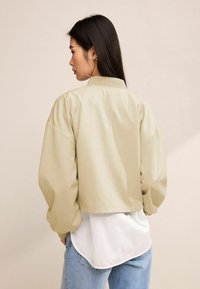 Beige cropped bomber dzseki gazdag ujjal, bordás gallérral és hátsó varrás részletekkel, fehér ing felett és világoskék farmerral viselve.