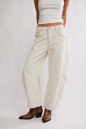 Kobieta w kremowych spodniach o szerokich nogawkach, białym prążkowanym topie na ramiączkach i brązowych skórzanych botkach, stojąca na jednolitym tle.