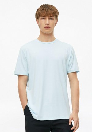 Młody mężczyzna o jasnobrązowych włosach, ubrany w gładki jasnoniebieski t-shirt z krótkim rękawem i czarne spodnie, stojący na białym tle.