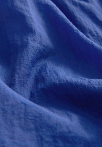 Sininen kangas, jossa on teksturoitu pinta, jossa on pehmeitä poimuja ja rypytyksiä, mikä korostaa sen kevyitä ja sileitä ominaisuuksia.