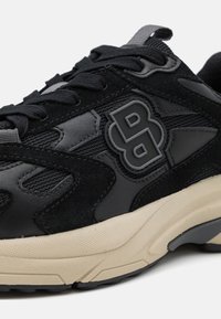 Sneaker de sport noir avec un mélange de suede et de matériaux synthétiques, semelle en caoutchouc texturé et un logo en relief bien visible.