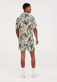Camicia a maniche corte con bottone e pantaloni corti coordinati, entrambi con motivi tropicali in verde e nero. Il tessuto ha una consistenza leggera e liscia.