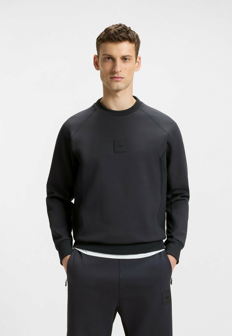 Herren schwarzer Pullover mit strukturierter Oberfläche, Rundhalsausschnitt, gerippten Bündchen und einem kleinen Logo-Patch auf der Vorderseite. Hat eine lockere Passform.