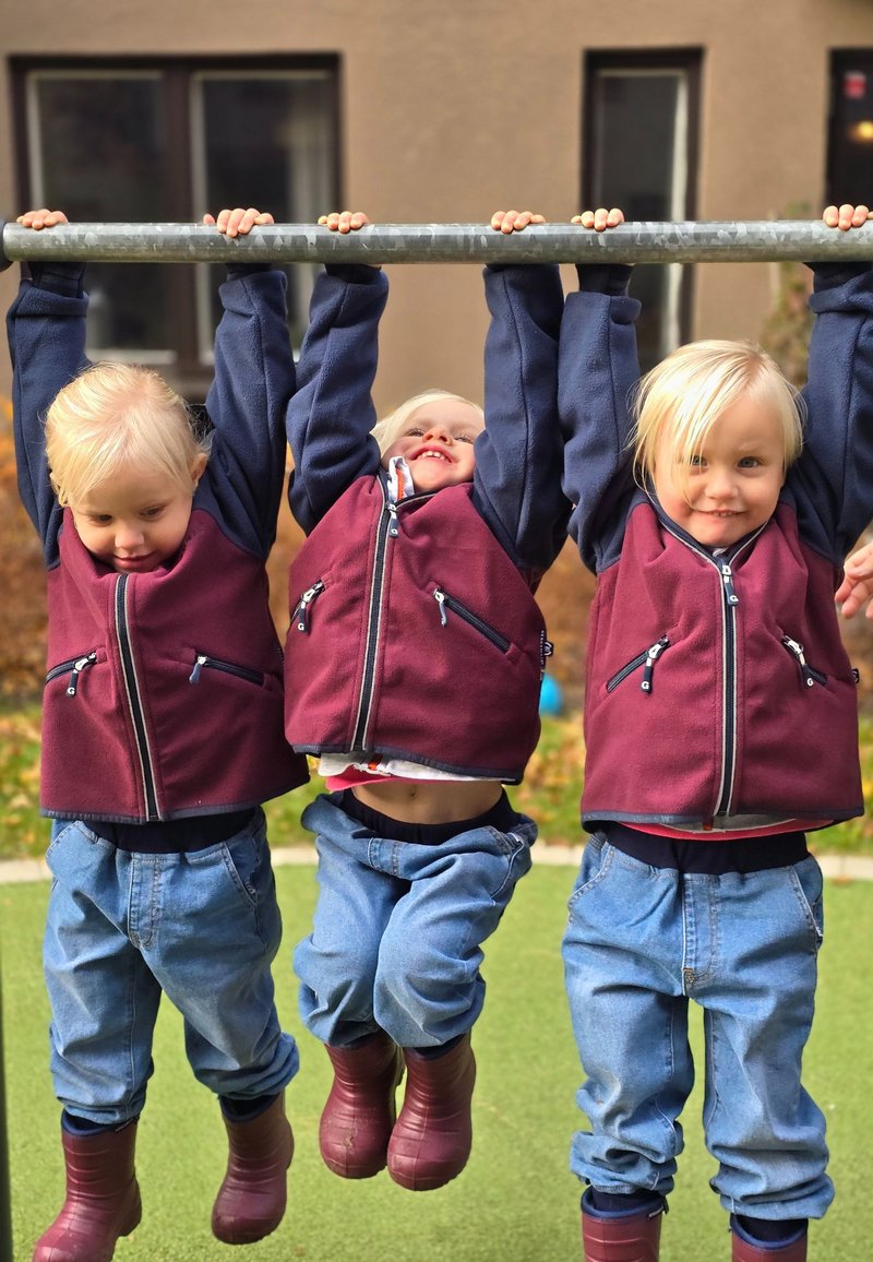 Tre barn hänger från en metallstång, iklädda vinröda zip-up västar med marinblå ärmar, blå jeans och bruna gummistövlar. Gräs i bakgrunden.