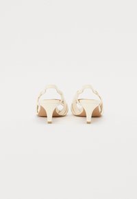 Beige High-Heelsandalen mit offenem Rücken, geschwungenen Riemen und einem schmalen Absatz. Aus glattem Material gefertigt und im minimalistischen Design.