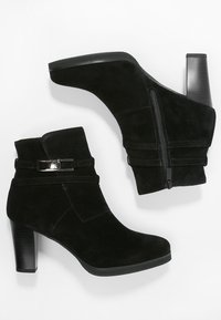 Bottines noires en suede avec un talon carré, fermeture éclair et deux sangles accentuées avec un matériel métallique. Texture lisse et bout pointu.