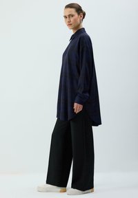 Marineblå ternet skjorte med knapper og lange ærmer, buet kant, båret over sorte bukser med vide ben og hvide sneakers.