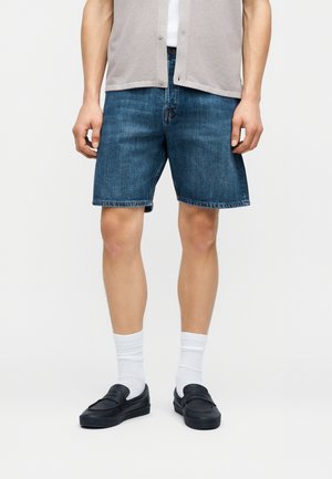 Hombre que lleva pantalones cortos de mezclilla azul hasta la mitad del muslo, calcetines blancos hasta la altura del tobillo, mocasines negros, una camisa blanca y un chaleco beige abotonado, posando frente a un fondo blanco.