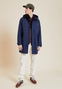 Marineblaue wasserdichte Jacke mit Kapuze, weißen Zugbändern, über ein bordeauxrotes Shirt geschichtet, kombiniert mit cremefarbenen Hosen und braunen Sneakern.