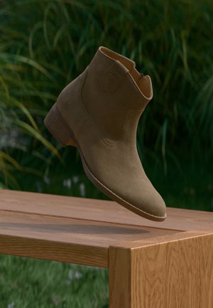 Ein brauner Wildleder-Knöchelstiefel schwebt über einer Holzbank mit verschwommenen grünen Pflanzen im Hintergrund.