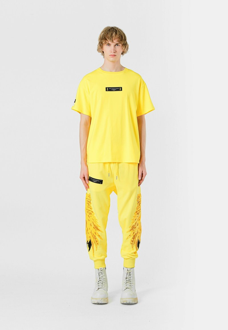 Camiseta de algodón amarilla de manga corta con una etiqueta negra, combinada con pantalones joggers a juego que presentan un diseño gráfico y detalles en negro.