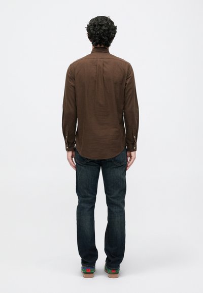 Hombre con cabello rizado oscuro, vistiendo una camisa de manga larga marrón, jeans oscuros y zapatillas de colores, de pie frente a un fondo blanco.