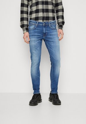 Mann trägt eine slim-fit blaue Jeans, ein schwarz-grau kariertes Hemd und schwarze Sneakers, steht vor einem schlichten hellen Hintergrund.