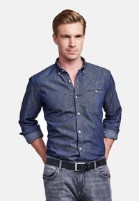 Denim shirt in deep blue, long sleeves rolled up, button-down collar, front pocket, subtle stitching details, paired with gray jeans and black belt.