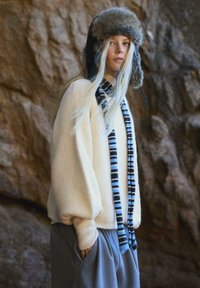 Krémový sveter s balónovými rukávmi, šedé teplákové nohavice a prúžkovaný čierny a modrý šál; čiapka s kožúškom a ušnými klapkami. Textúrovaný, neformálny outfit.