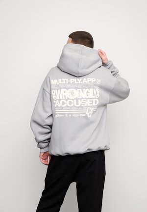 Person vendt væk bærer en lysegrå hættetrøje med stor hvid tekst på ryggen, der lyder "WRONGLY ACCUSED", og sorte bukser mod en ensfarvet baggrund.