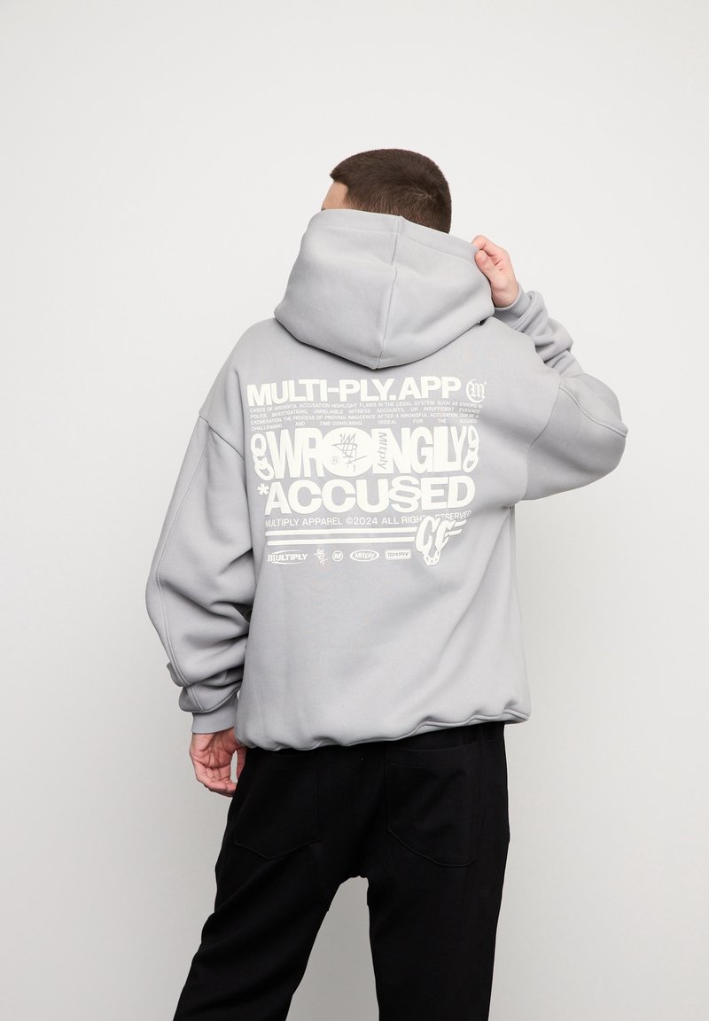 Personne de dos portant un sweat à capuche gris clair avec un grand texte blanc au dos indiquant "WRONGLY ACCUSED" et un pantalon noir sur un fond uni.