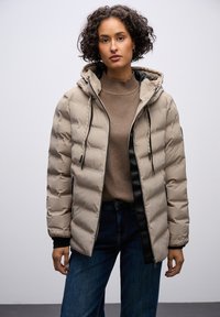 Beige dunjakke med hætte, struktureret overflade, lynlås foran og sorte detaljer. Bæres over en brun sweater og blå jeans.