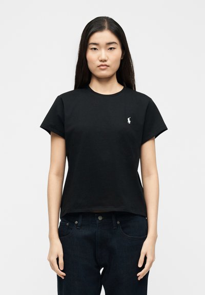 Γυναίκα με μακριά σκούρα μαλλιά, φοράει μαύρο κοντομάνικο μπλουζάκι με ένα μικρό λευκό λογότυπο στο στήθος και σκούρο τζιν, στέκεται μπροστά σε απλό φόντο.