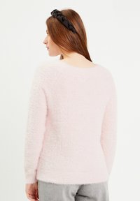 Zacht, roze, fluffy sweater met een geribbelde rand en ronde hals. Het model draagt een zwarte gedraaide hoofdband en toont het ontwerp aan de achterkant van de sweater.