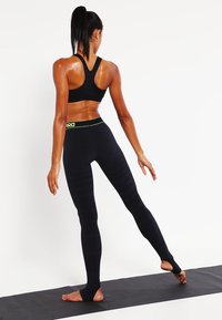 Černá sportovní podprsenka a legíny s vysokým pasem a neonově zeleným páskem. Legíny mají texturované vertikální pruhy. Stojí na jógové podložce.