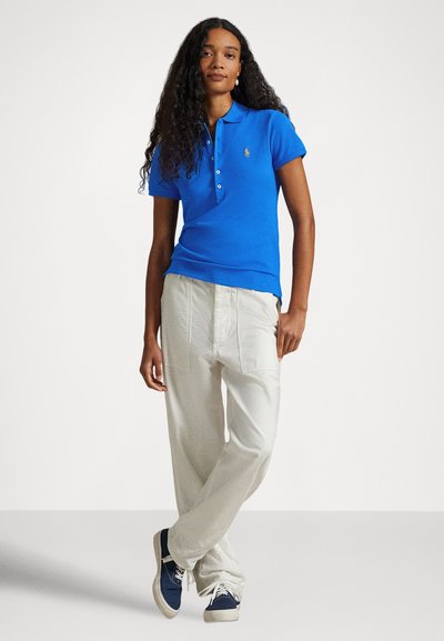 Μπλε βαμβακερό πόλο μπλουζάκι με κοντά μανίκια, κούμπωμα με τρία κουμπιά, συνδυασμένο με παντελόνι φαρδύ σε ανοιχτό μπεζ και μπλε αθλητικά παπούτσια.