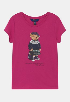Magenta t-shirt dziecięcy z grafiką misia w granatowym swetrze, w kratkę spódnicy, w sneakersach i czapce, trzymającego czarną torbę.