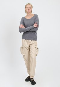 Maglione grigio a maglia con scollatura ampia, indossato con pantaloni cargo in ecopelle beige dotati di tasche frontali e polsini elasticizzati. Scarpe nere senza lacci.
