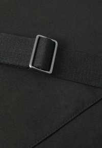 Tessuto nero con una finitura testurizzata, caratterizzato da un cinturino nero fissato da una fibbia metallica. Linee pulite ed elementi di design minimalisti.