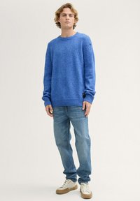 Blauer Strickpullover mit rundem Halsausschnitt, langen Ärmeln, gerippten Bündchen und Saum. Kombiniert mit hellblauen Jeans und beigen Sneakers.