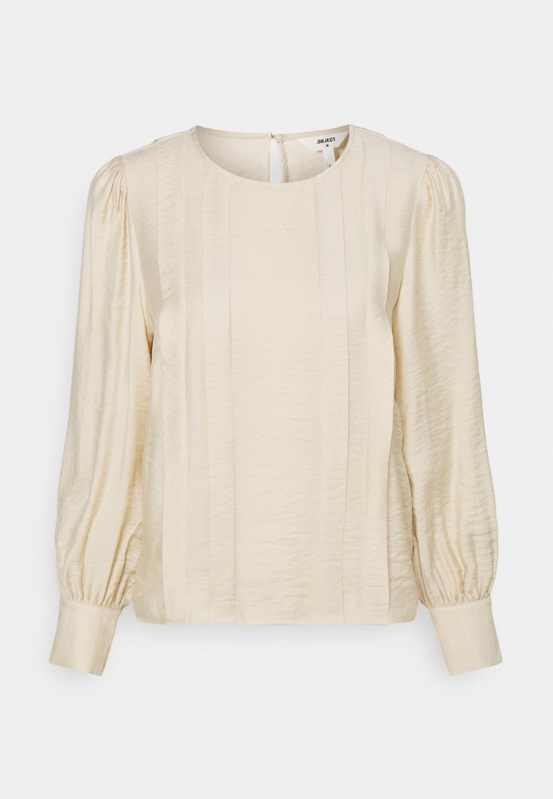 Cream-colored blouse with a round neckline, puffed sleeves, and vertical pleats. Soft, textured fabric and button closure at the back.