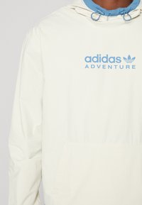 Ljusgrön hoodie-jacka med blå accenter, med en framficka och "adidas ADVENTURE"-logotyp. Slät textur, avslappnad design.