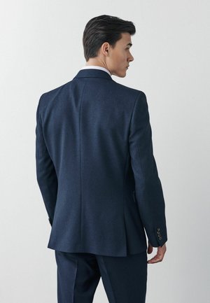 Chaqueta de traje azul marino a medida con un acabado texturizado, una abertura en la parte posterior y detalles de puño con cuatro botones. Diseño ajustado.