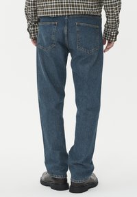 Modre kavbojke s ravnim rezom, izdelane iz denima z rahlo zbledelim videzom. Imajo dve zadnji žepni in kontrastno šivanje. Vidni so črni čevlji.