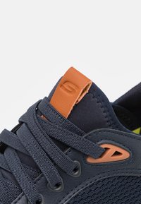 Zapatilla atlética en color navy con parte superior texturizada, detalle de cuero naranja en la lengüeta y cordones planos negros con diseño de malla para transpirabilidad.