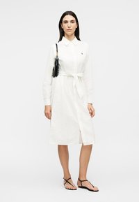 Femeie purtând o rochie cămașă albă cu centură, geantă neagră de umăr și sandale negre plate, stând în picioare pe un fundal alb simplu.